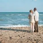 Older Couple Standing on Beach, Looking at Waves, His Arm Around Her