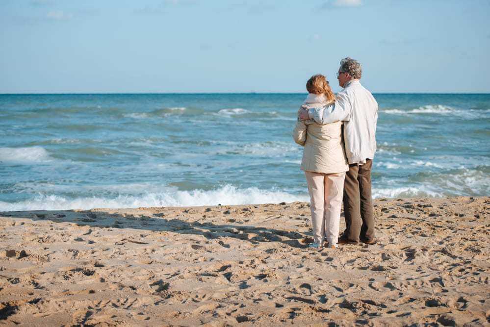 Older Couple Standing on Beach, Looking at Waves, His Arm Around Her